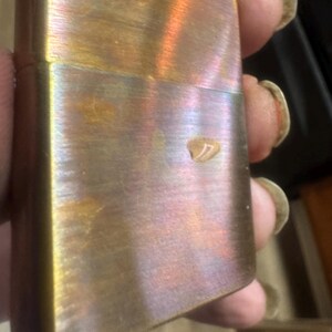 Pode incluir: Um isqueiro vintage de lat&atilde;o com um acabamento iridescente desgastado. A superf&iacute;cie do isqueiro exibe uma mistura de tons dourados, bronze e roxos, com uma pequena mancha mais clara. O isqueiro tem formato retangular com bordas arredondadas.