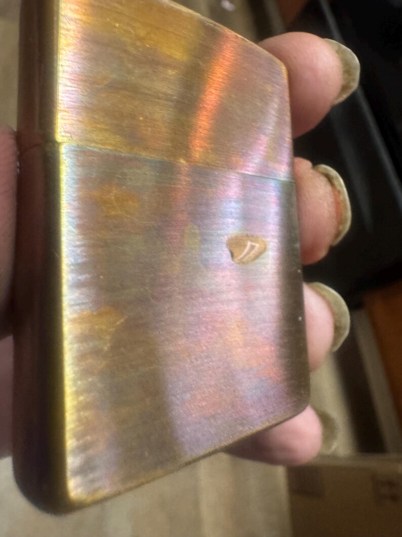 Pode incluir: Um isqueiro vintage de lat&atilde;o com um acabamento iridescente desgastado. A superf&iacute;cie do isqueiro exibe uma mistura de tons dourados, bronze e roxos, com uma pequena mancha mais clara. O isqueiro tem formato retangular com bordas arredondadas.