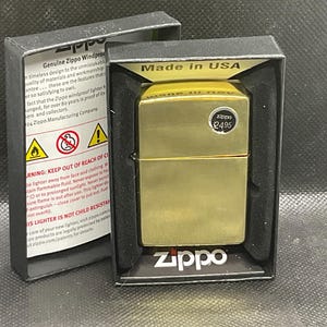以下が含まれることがあります： ブラッシュドメタル仕上げのゴールドトーンのZippo防風ライター。黒い段ボール製のディスプレイボックスに入っています。ボックスの内側には安全上の警告文とZippoのロゴがあり、ライターには円形の価格ステッカーが貼られています。