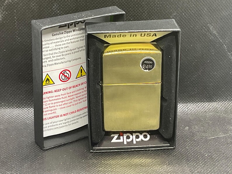 以下が含まれることがあります： ブラッシュドメタル仕上げのゴールドトーンのZippo防風ライター。黒い段ボール製のディスプレイボックスに入っています。ボックスの内側には安全上の警告文とZippoのロゴがあり、ライターには円形の価格ステッカーが貼られています。