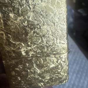 May include: A rectangular gold-colored metal lighter featuring a highly textured, crinkled surface pattern across its entire exterior. The lighter has a horizontal seam separating the lid from the main body, with a metallic finish that reflects light.