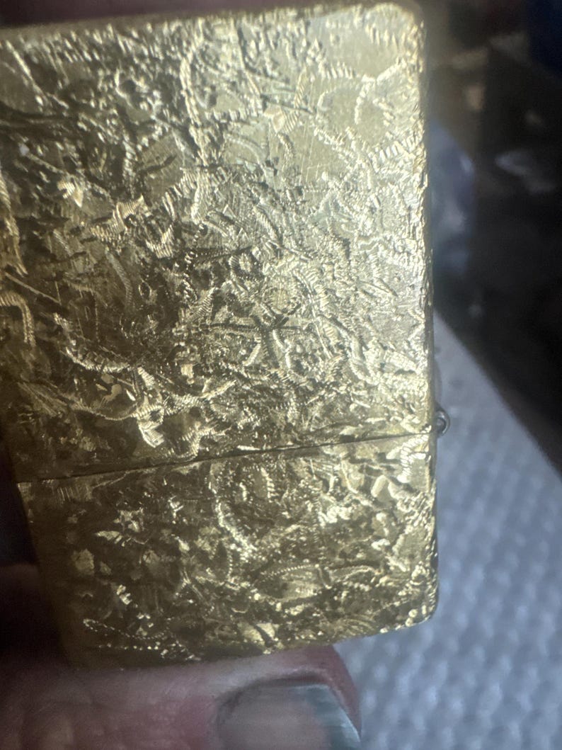 May include: A rectangular gold-colored metal lighter featuring a highly textured, crinkled surface pattern across its entire exterior. The lighter has a horizontal seam separating the lid from the main body, with a metallic finish that reflects light.