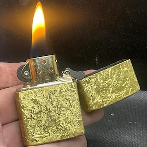 May include: A gold-colored, textured lighter with an open lid and a lit flame. The lighter is held in a hand, showcasing its intricate design. The flame is bright yellow and orange.