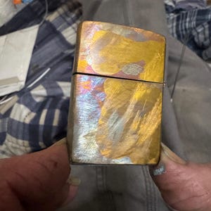 Pode incluir: Um isqueiro met&aacute;lico retangular com um acabamento iridescente colorido. A superf&iacute;cie do isqueiro exibe uma mistura de tons dourados, bronze e prata, criando uma apar&ecirc;ncia &uacute;nica e texturizada. A parte superior do isqueiro est&aacute; aberta, revelando o interior.