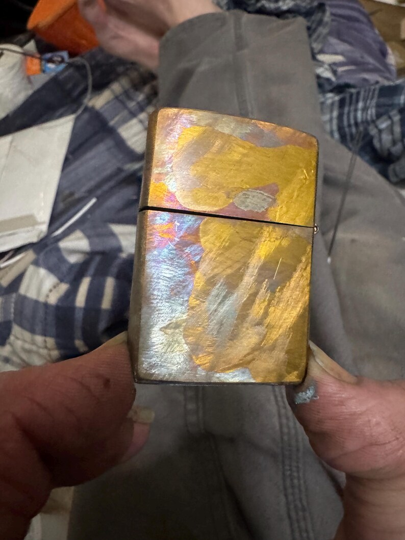 Pode incluir: Um isqueiro met&aacute;lico retangular com um acabamento iridescente colorido. A superf&iacute;cie do isqueiro exibe uma mistura de tons dourados, bronze e prata, criando uma apar&ecirc;ncia &uacute;nica e texturizada. A parte superior do isqueiro est&aacute; aberta, revelando o interior.