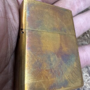 Pode incluir: Um isqueiro retangular de lat&atilde;o com tampa articulada. O isqueiro tem uma apar&ecirc;ncia desgastada e envelhecida com uma mistura de tons dourados, castanhos e toques de azul e roxo. A superf&iacute;cie mostra sinais de uso e desgaste.