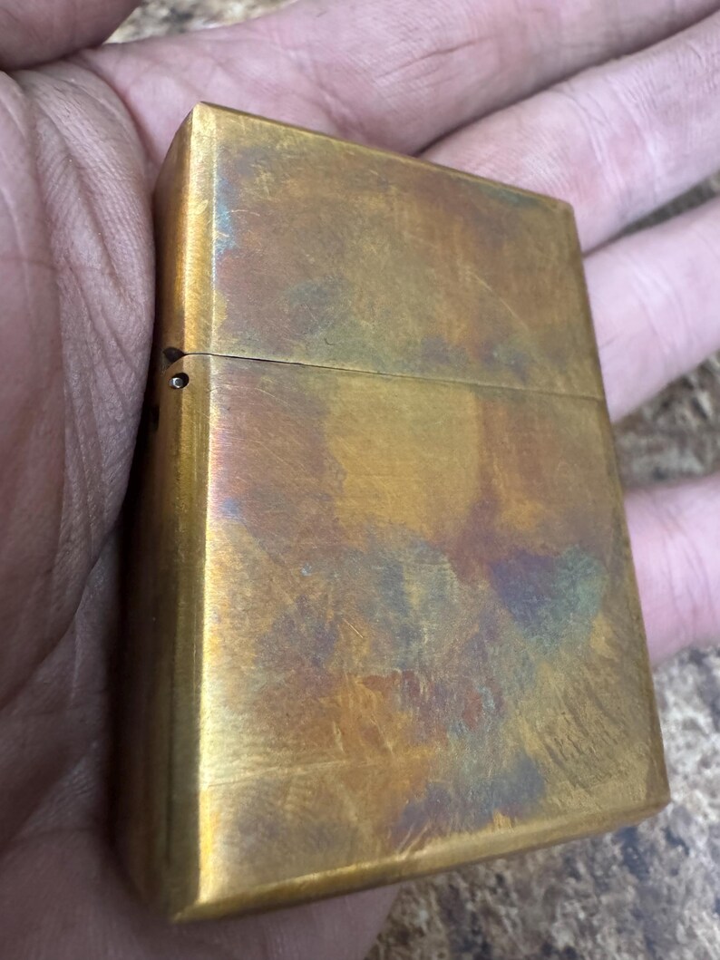 Pode incluir: Um isqueiro retangular de lat&atilde;o com tampa articulada. O isqueiro tem uma apar&ecirc;ncia desgastada e envelhecida com uma mistura de tons dourados, castanhos e toques de azul e roxo. A superf&iacute;cie mostra sinais de uso e desgaste.