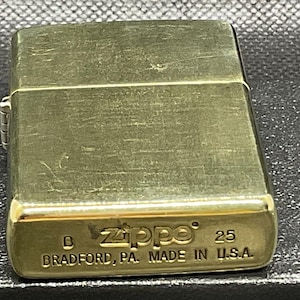 以下が含まれることがあります： ヒンジ付きの蓋が付いた長方形のゴールドトーンの金属製ライター。底面には、Zippoのエンボス加工されたテキスト、数字の25、およびBRADFORD, PA. MADE IN U.S.A.という刻印があり、底面の左側には文字Bが確認できます。