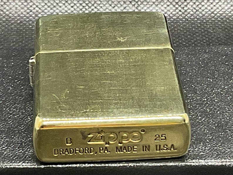 以下が含まれることがあります： ヒンジ付きの蓋が付いた長方形のゴールドトーンの金属製ライター。底面には、Zippoのエンボス加工されたテキスト、数字の25、およびBRADFORD, PA. MADE IN U.S.A.という刻印があり、底面の左側には文字Bが確認できます。