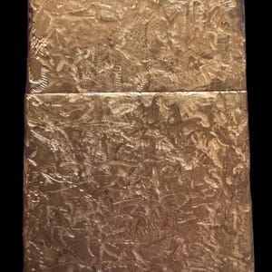 May include: A rectangular gold-colored metal lighter featuring a heavily textured, hammered surface with deep, irregular indentations across the entire exterior. The lighter has a horizontal seam separating the lid from the main body section.