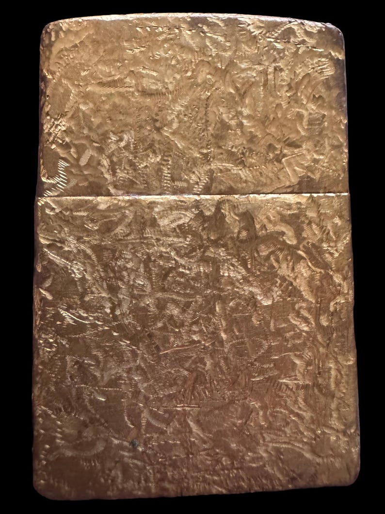 May include: A rectangular gold-colored metal lighter featuring a heavily textured, hammered surface with deep, irregular indentations across the entire exterior. The lighter has a horizontal seam separating the lid from the main body section.