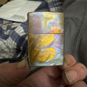 Pode incluir: Um isqueiro met&aacute;lico retangular com um acabamento iridescente. A superf&iacute;cie do isqueiro exibe uma mistura de tons dourados, azuis e roxos, criando um efeito visual marcante. O isqueiro &eacute; segurado em uma m&atilde;o, sugerindo sua natureza port&aacute;til.