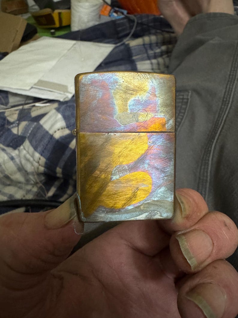 Pode incluir: Um isqueiro met&aacute;lico retangular com um acabamento iridescente. A superf&iacute;cie do isqueiro exibe uma mistura de tons dourados, azuis e roxos, criando um efeito visual marcante. O isqueiro &eacute; segurado em uma m&atilde;o, sugerindo sua natureza port&aacute;til.