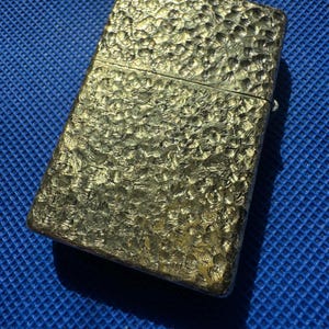 May include: A rectangular metal lighter with a gold-toned finish and a distinct hammered, dimpled texture across its entire surface. The lighter features a hinged lid design with a horizontal seam separating the top cap from the main body.
