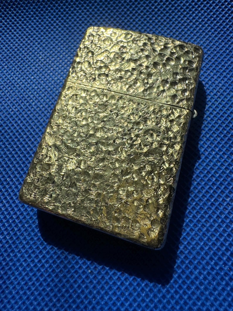 May include: A rectangular metal lighter with a gold-toned finish and a distinct hammered, dimpled texture across its entire surface. The lighter features a hinged lid design with a horizontal seam separating the top cap from the main body.