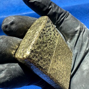 May include: A rectangular gold-colored metal lighter featuring a heavily hammered, textured surface across its entire exterior. The lighter has a distinct horizontal seam separating the lid from the base, with a metallic finish that reflects light.