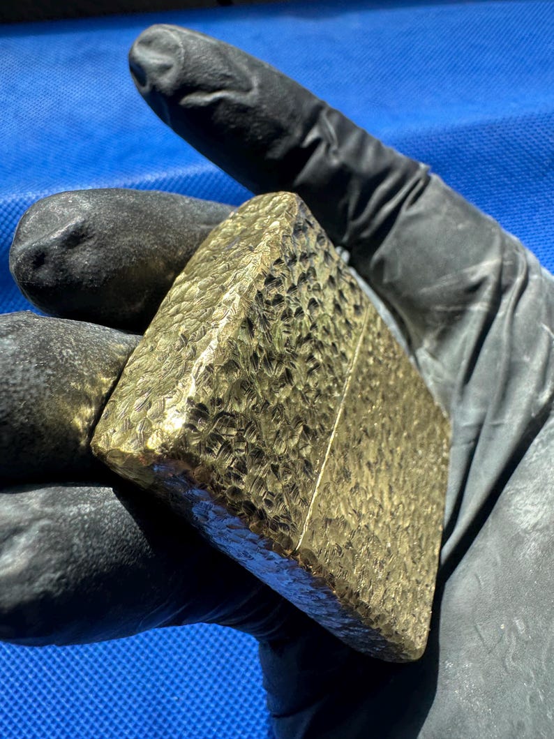 May include: A rectangular gold-colored metal lighter featuring a heavily hammered, textured surface across its entire exterior. The lighter has a distinct horizontal seam separating the lid from the base, with a metallic finish that reflects light.
