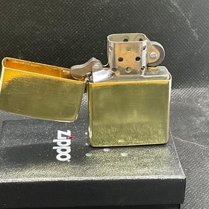 以下が含まれることがあります： 光沢のある仕上げのゴールドトーンの金属製フリップトップライター。蓋が開いており、銀色の内部チムニーとフリントホイール機構が見えます。ライターは、Zippoのロゴが印刷された黒い長方形の箱の上に置かれています。
