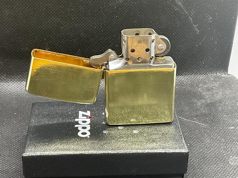 以下が含まれることがあります： 光沢のある仕上げのゴールドトーンの金属製フリップトップライター。蓋が開いており、銀色の内部チムニーとフリントホイール機構が見えます。ライターは、Zippoのロゴが印刷された黒い長方形の箱の上に置かれています。