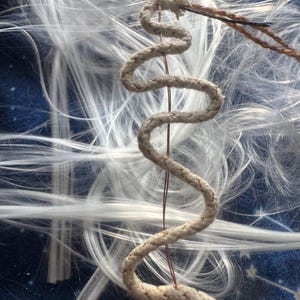 May include: Close-up of a cream-colored, coiled rope with red stitching, held by a needle and thread. White, wispy fibers surround the rope against a dark blue, patterned background. The rope is the focal point.