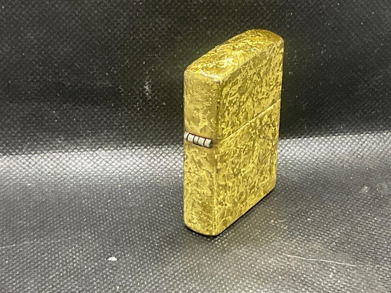 May include: A rectangular metal lighter with a textured, mottled gold-tone finish. The lighter features a hinged lid and a visible silver-colored metal hinge mechanism on the side, showcasing a metallic surface with an irregular, embossed pattern.