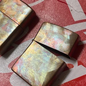 Pode incluir: Dois isqueiros iridescentes com acabamento perolado. Um isqueiro est&aacute; aberto, revelando o interior. Os isqueiros t&ecirc;m formato retangular com bordas arredondadas e uma moldura bronze. O fundo &eacute; vermelho e branco.