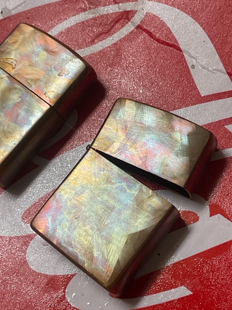 Pode incluir: Dois isqueiros iridescentes com acabamento perolado. Um isqueiro est&aacute; aberto, revelando o interior. Os isqueiros t&ecirc;m formato retangular com bordas arredondadas e uma moldura bronze. O fundo &eacute; vermelho e branco.