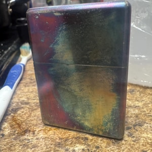 Pode incluir: Um isqueiro retangular de metal com um acabamento iridescente colorido. A superf&iacute;cie do isqueiro exibe uma mistura de cores, incluindo vermelho, azul, verde e amarelo, criando uma apar&ecirc;ncia art&iacute;stica &uacute;nica. O isqueiro est&aacute; em p&eacute;.