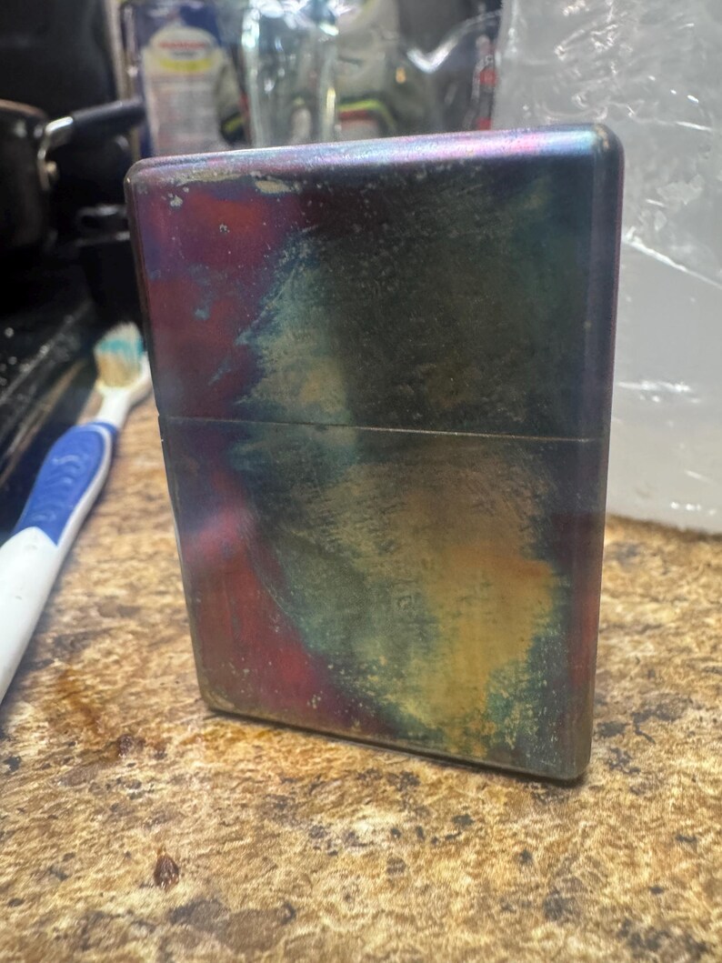 Pode incluir: Um isqueiro retangular de metal com um acabamento iridescente colorido. A superf&iacute;cie do isqueiro exibe uma mistura de cores, incluindo vermelho, azul, verde e amarelo, criando uma apar&ecirc;ncia art&iacute;stica &uacute;nica. O isqueiro est&aacute; em p&eacute;.