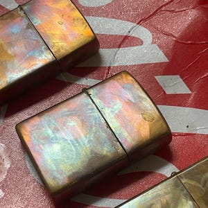 Pode incluir: Close-up de tr&ecirc;s isqueiros de estilo vintage com um acabamento met&aacute;lico iridescente. Os isqueiros t&ecirc;m formato retangular com tampa articulada. As cores variam do bronze ao dourado, com toques de azul e roxo. O fundo &eacute; uma superf&iacute;cie vermelha e branca.