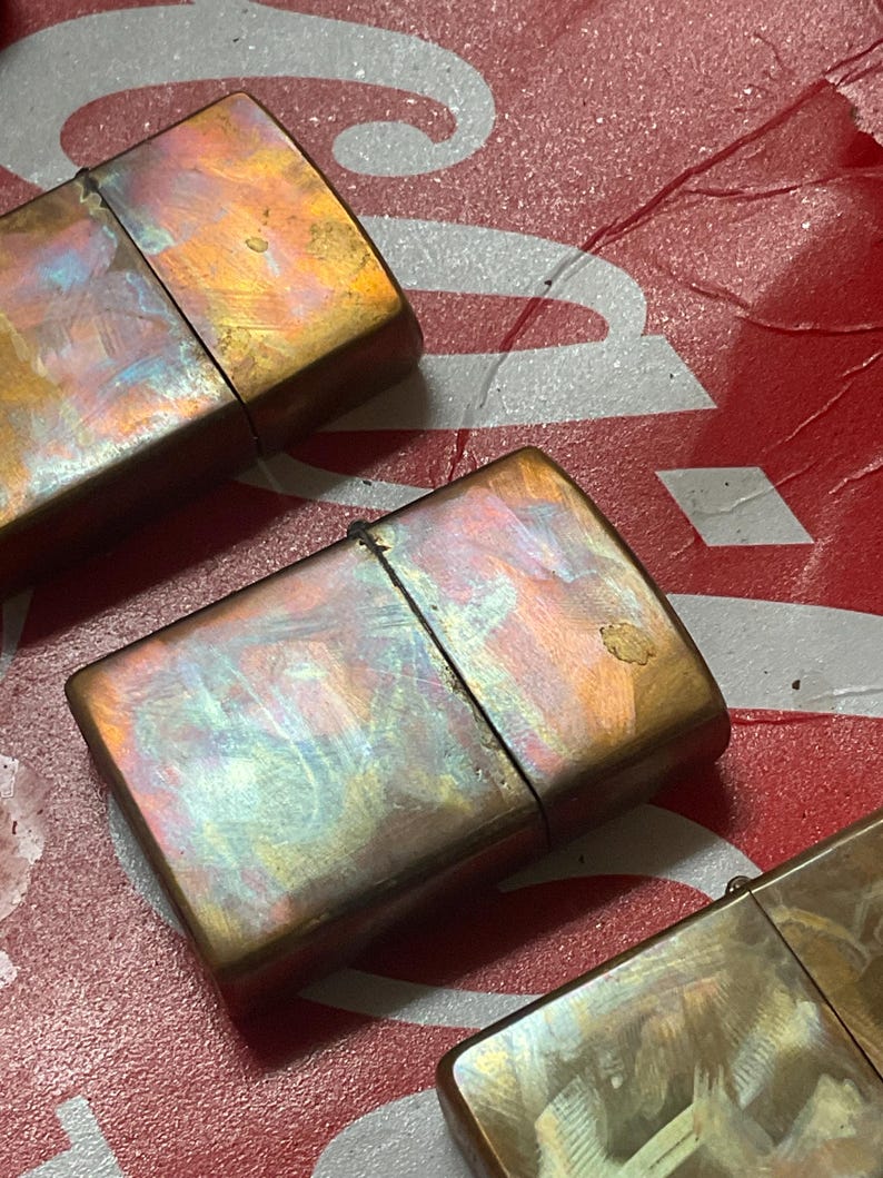 Pode incluir: Close-up de tr&ecirc;s isqueiros de estilo vintage com um acabamento met&aacute;lico iridescente. Os isqueiros t&ecirc;m formato retangular com tampa articulada. As cores variam do bronze ao dourado, com toques de azul e roxo. O fundo &eacute; uma superf&iacute;cie vermelha e branca.