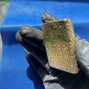 May include: A rectangular gold-toned metal lighter featuring a highly textured, hammered surface finish. The lighter has a hinged lid design and a metallic sheen that reflects light across its uneven, dimpled exterior pattern.