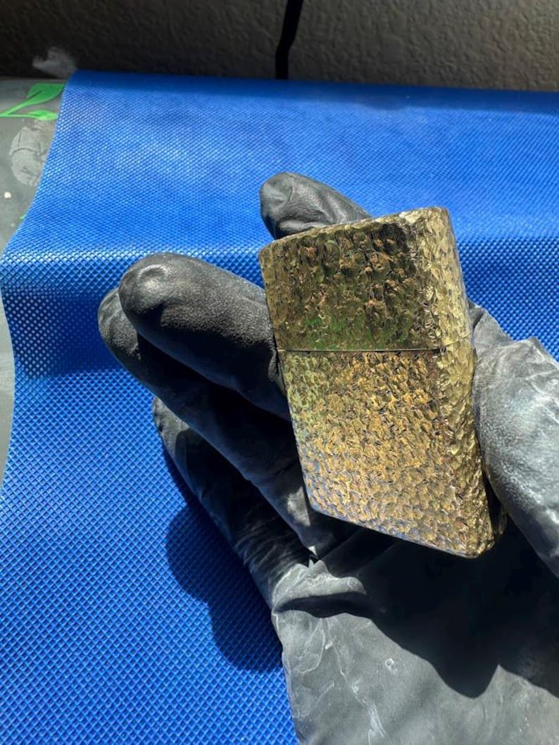 May include: A rectangular gold-toned metal lighter featuring a highly textured, hammered surface finish. The lighter has a hinged lid design and a metallic sheen that reflects light across its uneven, dimpled exterior pattern.
