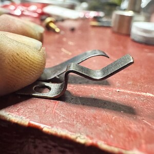 May include: A small, silver-colored metal spring clip featuring a flat base with a circular hole and a bent, angular arm extending upward. The component has a metallic finish and a distinct, multi-angled shape designed for mechanical tension.