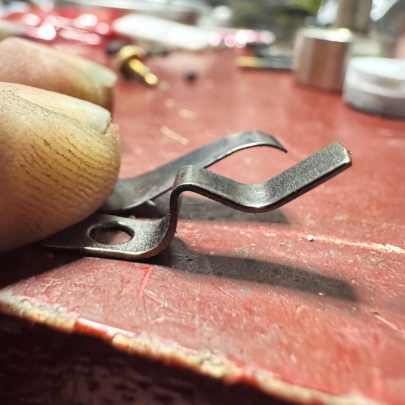 May include: A small, silver-colored metal spring clip featuring a flat base with a circular hole and a bent, angular arm extending upward. The component has a metallic finish and a distinct, multi-angled shape designed for mechanical tension.
