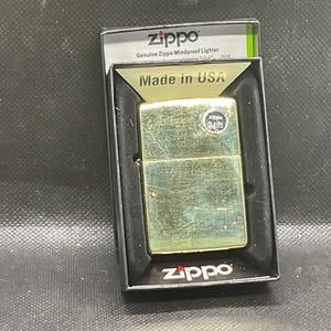 以下が含まれることがあります： 黒いディスプレイボックスに入ったゴールドトーンのZippo防風ライター。ライターは長方形の金属製ボディで、2495と書かれた小さな円形のステッカーが貼られています。パッケージにはZippo、Genuine Zippo Windproof Lighter、Made in USAという文字が記載されています。