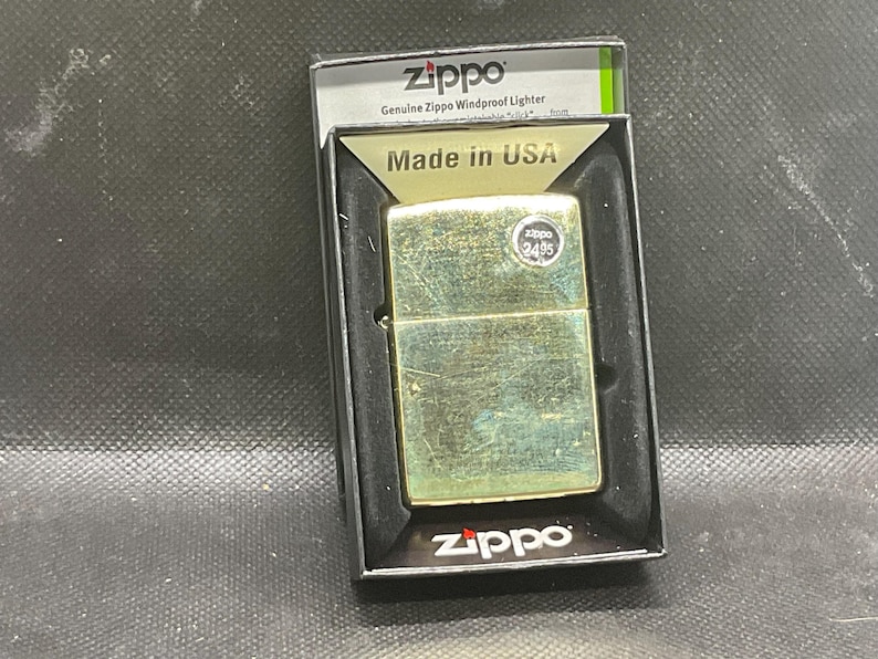 以下が含まれることがあります： 黒いディスプレイボックスに入ったゴールドトーンのZippo防風ライター。ライターは長方形の金属製ボディで、2495と書かれた小さな円形のステッカーが貼られています。パッケージにはZippo、Genuine Zippo Windproof Lighter、Made in USAという文字が記載されています。