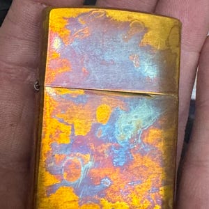 Pode incluir: Um isqueiro retangular cor de lat&atilde;o com um acabamento iridescente envelhecido. A superf&iacute;cie do isqueiro exibe uma mistura de tons dourados, azuis e laranja, criando uma apar&ecirc;ncia texturizada &uacute;nica. O isqueiro &eacute; segurado em uma m&atilde;o.