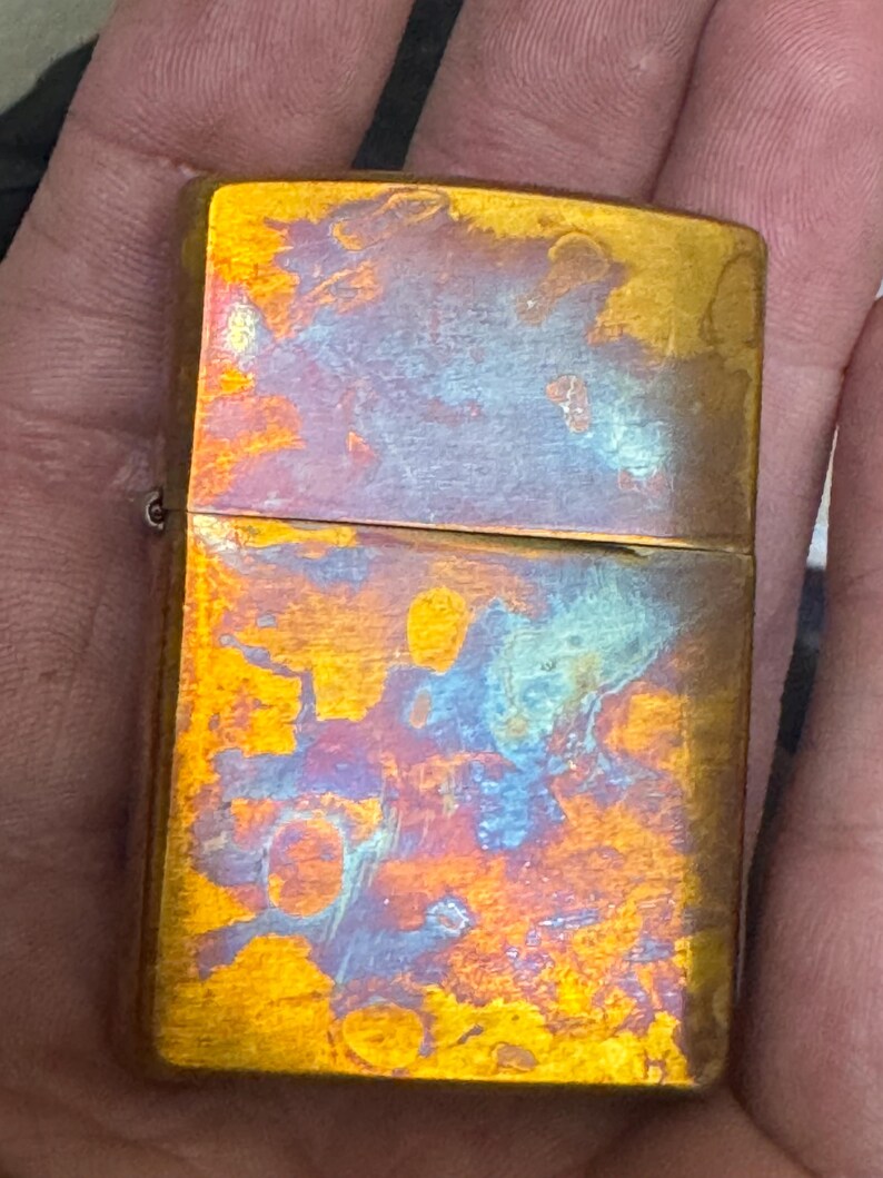 Pode incluir: Um isqueiro retangular cor de lat&atilde;o com um acabamento iridescente envelhecido. A superf&iacute;cie do isqueiro exibe uma mistura de tons dourados, azuis e laranja, criando uma apar&ecirc;ncia texturizada &uacute;nica. O isqueiro &eacute; segurado em uma m&atilde;o.