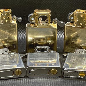May include: A collection of six metal lighter inserts arranged in two rows. The top row features three gold-toned brass inserts, while the bottom row consists of three silver-toned steel inserts. Each insert displays engraved text including Zippo branding.