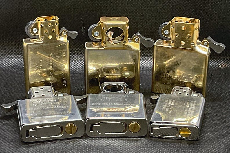 May include: A collection of six metal lighter inserts arranged in two rows. The top row features three gold-toned brass inserts, while the bottom row consists of three silver-toned steel inserts. Each insert displays engraved text including Zippo branding.