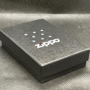 以下が含まれることがあります： 表面に質感のある長方形の黒い厚紙製ギフトボックス。箱の上面中央には白いZippoのロゴが印刷されており、その上部には8つの小さな水色の点が正方形のパターンで配置されています。