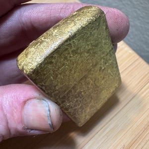 May include: A rectangular gold-colored metal lighter featuring a textured, mottled surface finish. The lighter has a hinged lid design with a visible horizontal seam separating the top cap from the main body, showcasing a metallic, reflective appearance.