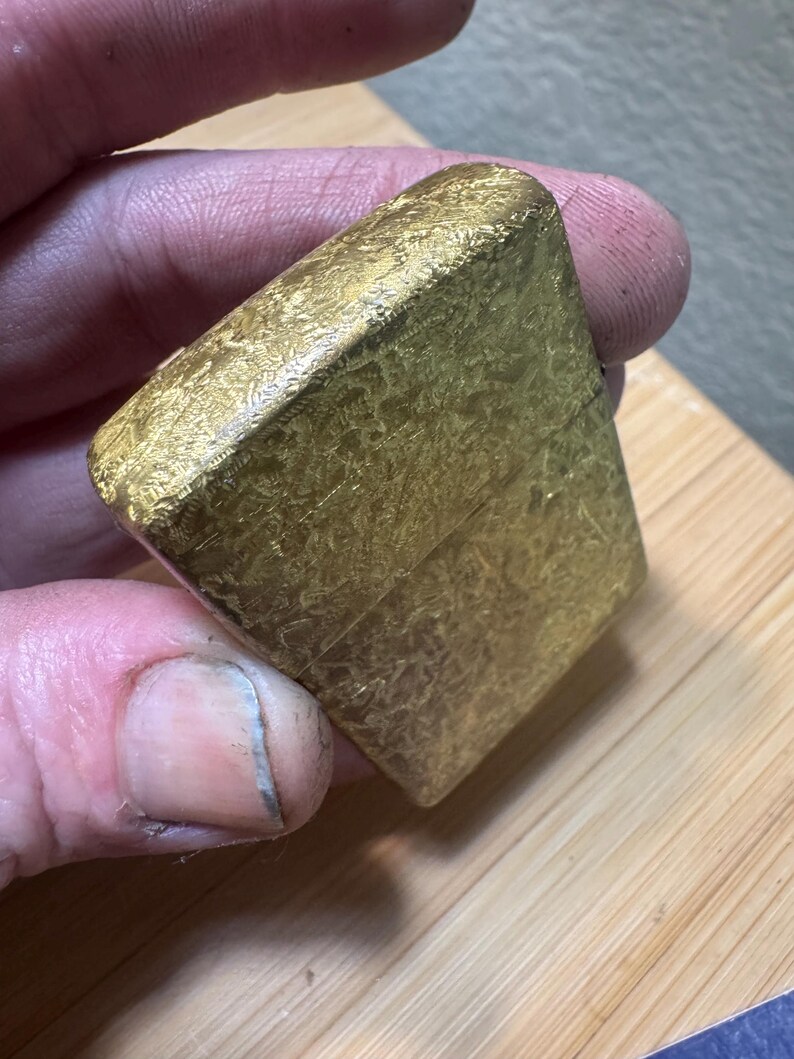 May include: A rectangular gold-colored metal lighter featuring a textured, mottled surface finish. The lighter has a hinged lid design with a visible horizontal seam separating the top cap from the main body, showcasing a metallic, reflective appearance.