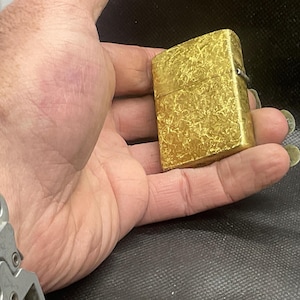 May include: A rectangular gold-colored metal lighter featuring a textured, mottled surface pattern. The lighter has a hinged lid and a metallic finish, showing a distinct cross-hatched or scratched design across its entire exterior surface.