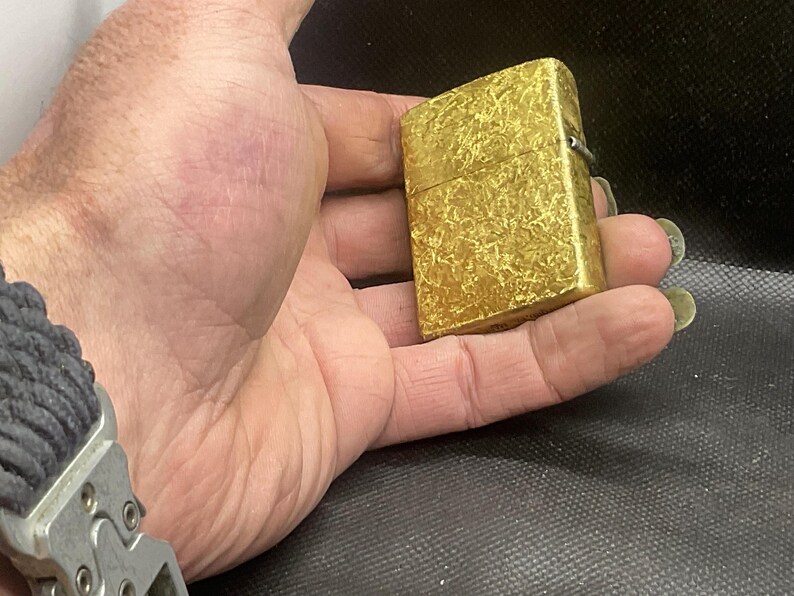 May include: A rectangular gold-colored metal lighter featuring a textured, mottled surface pattern. The lighter has a hinged lid and a metallic finish, showing a distinct cross-hatched or scratched design across its entire exterior surface.