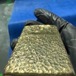 May include: A rectangular metal lighter with a textured, hammered gold-tone finish. The surface features an uneven, pebbled pattern that catches the light, creating highlights and shadows across the metallic casing of the flip-top lighter.