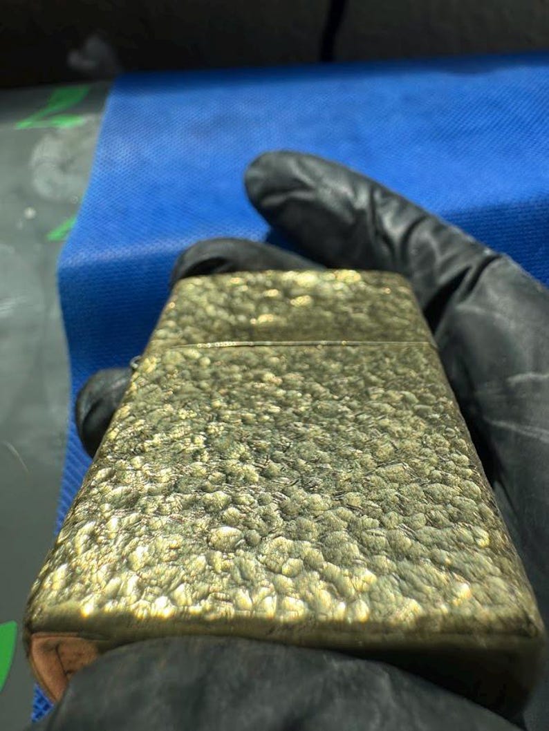 May include: A rectangular metal lighter with a textured, hammered gold-tone finish. The surface features an uneven, pebbled pattern that catches the light, creating highlights and shadows across the metallic casing of the flip-top lighter.