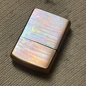 Pode incluir: Um isqueiro de estilo vintage com acabamento met&aacute;lico. A superf&iacute;cie do isqueiro exibe uma mistura de cores, incluindo dourado, roxo e bronze. A forma retangular e a tampa articulada s&atilde;o vis&iacute;veis. O isqueiro est&aacute; sobre uma superf&iacute;cie castanha texturizada.