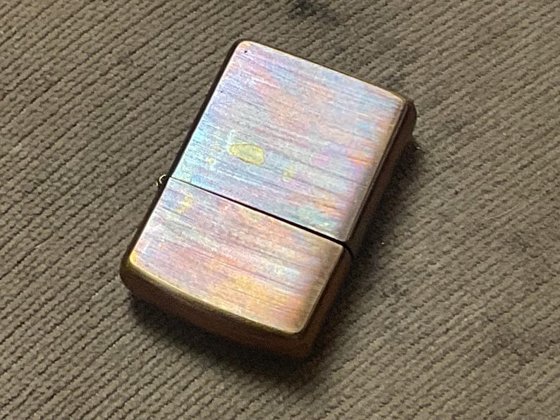 Pode incluir: Um isqueiro de estilo vintage com acabamento met&aacute;lico. A superf&iacute;cie do isqueiro exibe uma mistura de cores, incluindo dourado, roxo e bronze. A forma retangular e a tampa articulada s&atilde;o vis&iacute;veis. O isqueiro est&aacute; sobre uma superf&iacute;cie castanha texturizada.